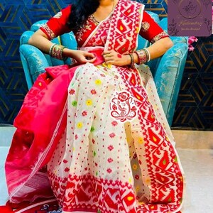 May include: A red and white patterned saree with a red blouse. The saree features a detailed design with floral and geometric patterns. The blouse has intricate embroidery. The saree is draped over a person sitting in a blue chair.
