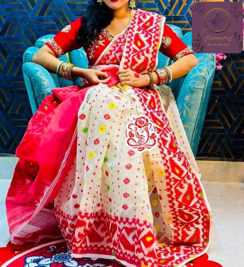 May include: A red and white patterned saree with a red blouse. The saree features a detailed design with floral and geometric patterns. The blouse has intricate embroidery. The saree is draped over a person sitting in a blue chair.