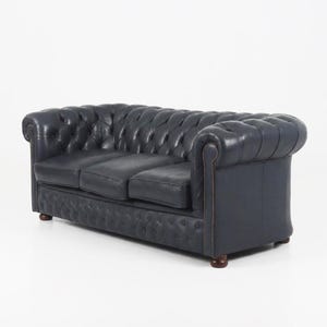 Op de afbeelding: Een donkerblauwe Chesterfield-bank met een gecapitonneerde rugleuning en zitting. De bank heeft opgerolde armleuningen en wordt ondersteund door korte, donkere houten poten. De bekleding is een glad, leerachtig materiaal. De bank is een driezitter.