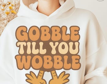 May include: White hooded sweatshirt with the text "GOBBLE TILL YOU WOBBLE" in brown and tan retro font. The design includes two cartoon turkey hands. A fun, festive design for Thanksgiving.