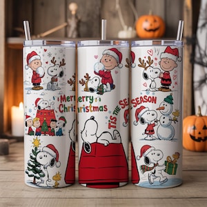 May include: White stainless steel tumbler featuring Snoopy and Charlie Brown in Christmas scenes. The tumbler has a straw and is decorated with holiday graphics and the text "Merry Christmas" and "Tis the Season".