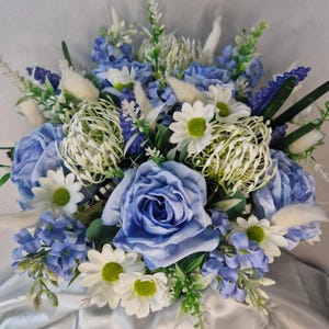 May include: A floral arrangement featuring light blue roses, white daisies with green centres, purple lavender-like sprigs, and white feathery plumes. The bouquet includes various green foliage and stems, creating a dense, rounded display of mixed artificial flowers in shades of blue, white, and green.