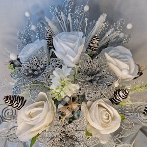 May include: A decorative floral arrangement featuring white roses, silver glittered poinsettias, and small pinecones. The bouquet includes white berry accents, silver wire swirls, and a small gift box wrapped in black and white buffalo check fabric.