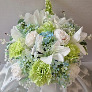 May include: A floral arrangement featuring white lilies, pale green carnations, and small light blue blossoms. The bouquet includes delicate white butterfly accents and green foliage, all gathered together in a rounded, dense display of artificial flowers.