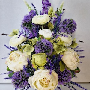 May include: A floral arrangement featuring cream-coloured peonies and ranunculus flowers interspersed with vibrant purple globe thistles, lavender sprigs, and light green foliage. The bouquet is structured in a vertical, tapered display.