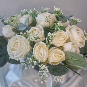 May include: A floral arrangement featuring multiple cream-coloured roses interspersed with small white buds and sage green eucalyptus leaves. The bouquet is densely packed with layered petals and foliage, creating a rounded, full appearance.