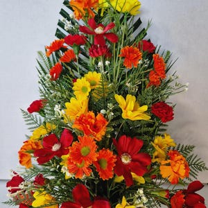 May include: A triangular floral arrangement featuring a vibrant mix of red, orange, and yellow artificial flowers. The display includes various daisy-like blooms, carnations, and green fern-like foliage, all arranged in a tall, pointed, vertical shape.