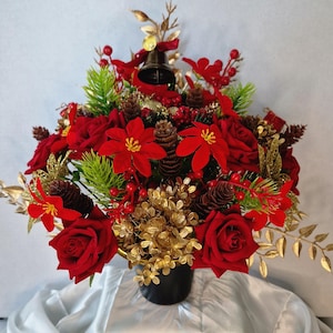 May include: A festive floral arrangement in a black container featuring deep red roses, bright red poinsettias, green pine sprigs, brown pinecones, gold-toned foliage, red berries, and a small metallic bell at the top centre of the display.