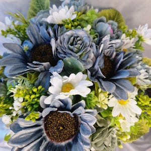 May include: A floral arrangement featuring dusty blue sunflowers with dark brown centres, white daisies with yellow centres, and small green sprigs. The bouquet includes various blue and green foliage, creating a dense, layered composition of artificial flowers with varied petal textures and shades.