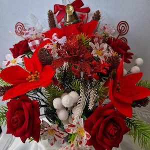 May include: A floral arrangement featuring red poinsettias, red roses, white berries, pinecones, and green pine needles. A gold-coloured bell with a red ribbon bow sits at the top. The display includes small white flowers with red speckles and red swirls.