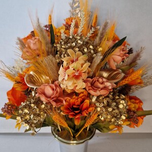 May include: A floral arrangement in a silver-toned metal vase featuring orange, peach, and gold-coloured artificial flowers, wheat stalks, and metallic gold foliage. The bouquet includes roses, chrysanthemums, and various dried-style grasses.