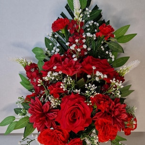 May include: A floral arrangement featuring deep red roses, carnations, and dahlias accented with small white baby's breath flowers and green foliage. The bouquet is shaped in a vertical, pointed design with dark green leaves at the top.