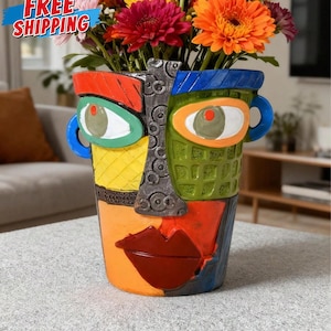 May include: A colorful, abstract face-shaped vase filled with vibrant flowers. The vase features bold colors like orange, blue, yellow, and red, with a textured design. The flowers include orange, pink, and yellow gerbera daisies, creating a striking floral arrangement.