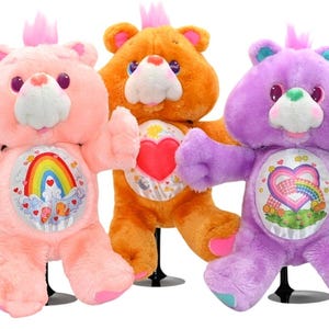 May include: Three plush Care Bears in pink, orange, and purple. Each bear has a unique belly badge with a rainbow, heart, or other design. They have a small crown on their heads and are standing on black stands.