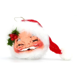 May include: A vintage Christmas decoration in the shape of Santa Claus's head. The ornament has a rosy-cheeked face, closed eyes, a white moustache, and a red hat with a white bobble. Holly and red berries decorate the side of the face.