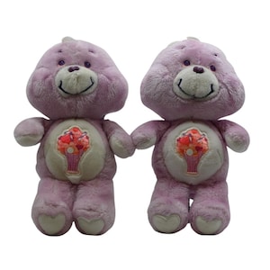May include: Two lavender plush bears with smiling faces and heart-shaped feet. Each bear has a belly emblem of a pink ice cream sundae in a purple cup. The bears are holding hands.