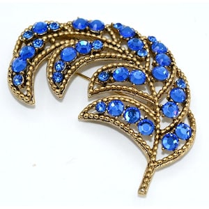 Op de afbeelding: Een vintage-stijl broche in de vorm van een veer. De broche heeft een goudkleurig metalen frame en is versierd met talrijke ronde, briljante blauwe strass-steentjes. Het ontwerp is ingewikkeld en gedetailleerd, met een klassieke, elegante uitstraling.