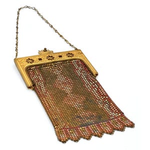 May include: An antique gold-toned metal mesh purse with a chain handle. The purse features a decorative frame with floral accents and a patterned mesh body in shades of red, brown, and gold. The bottom edge has a scalloped design.