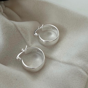 May include: A pair of silver hoop earrings with a shiny finish. The earrings have a rounded shape with a thick band and a hinged clasp. They are presented on a soft, light-coloured fabric.