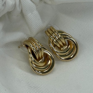 May include: A pair of gold-coloured earrings. Each earring has an oval design with multiple linked loops. The earrings have a post backing and are displayed on a white surface.