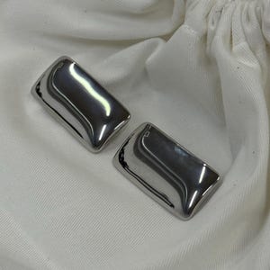 Op de afbeelding: Twee gepolijste, rechthoekige metalen objecten met gebogen randen. De objecten zijn reflecterend, zilverkleurig en liggen op een witte stoffen ondergrond. De items lijken decoratieve of functionele hardware te zijn.