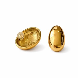 May include: A pair of gold-colored stud earrings. One earring is shown with the post and back, the other is a solid oval shape. The earrings have a shiny, reflective surface and a smooth finish. The earrings are set against a white background.