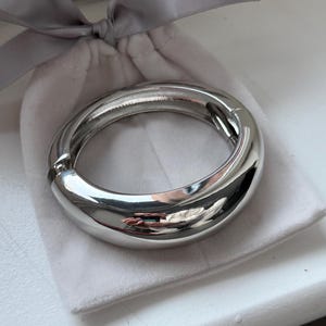 May include: A polished, silver-tone bangle bracelet with a smooth, rounded design. The bracelet sits on a white, velvet pouch, secured with a grey ribbon. The bracelet's surface reflects the surroundings.