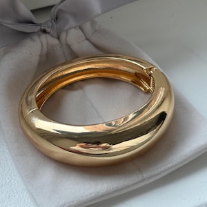 May include: A gold-tone, chunky bracelet with a hinged clasp. The bracelet is smooth and reflective, resting on a soft, white pouch. The bracelet's design is a thick, rounded band, suggesting a bold, modern style. The clasp is visible, indicating ease of wear.