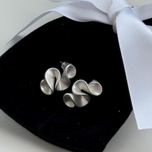 Puede incluir: Pendientes de botón plateados con un diseño único y fluido, que se asemeja a una cinta o una ola estilizada. Los pendientes se muestran sobre una bolsa de terciopelo negro, con una cinta blanca en el fondo.