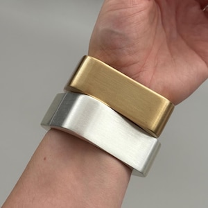 Könnte beinhalten: Zwei Metallarmbänder, eines goldfarben und eines silberfarben, sind an einem Handgelenk gestapelt. Die Armbänder haben eine rechteckige Form mit abgerundeten Kanten und einem gebürsteten Metall-Finish. Die Armbänder sind etwa 5 cm breit.