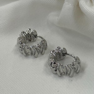 May include: A pair of silver hoop earrings. Each earring has a textured, ridged design around the hoop. The earrings are presented on a white, textured surface.