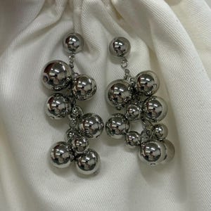 May include: A pair of silver dangle earrings. Each earring has multiple shiny, spherical beads of varying sizes, suspended from a small chain. The earrings reflect light, creating a bright, metallic appearance. The design is modern and eye-catching.