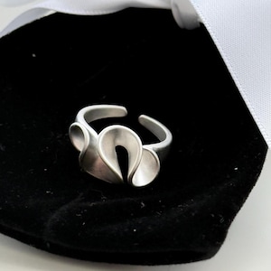 May include: A silver ring with an unusual, open design. The ring has three curved, petal-like shapes, giving it an abstract floral or ribbon-like look. The adjustable band allows for a comfortable fit.