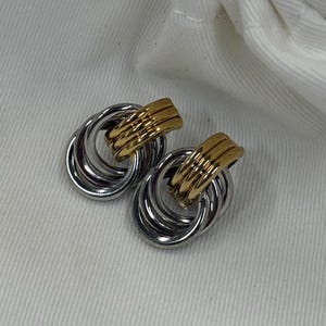 May include: A pair of silver-tone earrings with a gold-tone accent. Each earring features three interlocking rings with a gold-tone bar at the top. The earrings are displayed on a white surface.