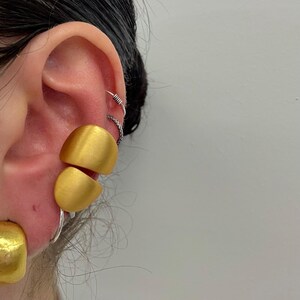 Peut inclure: Gros plan sur une oreille ornée de plusieurs boucles d'oreilles. Les boucles d'oreilles principales sont grandes, de couleur or, et ont un design géométrique arrondi. Des créoles et des boucles d'oreilles hélix argentées ajoutent du contraste.