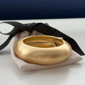 Könnte beinhalten: Ein goldfarbenes, breites Armreif-Armband mit gebürsteter Oberfläche, das auf einem kleinen, hellfarbigen Beutel liegt. Ein schwarzes Band ist um den Beutel gebunden. Das Armband hat ein glattes, abgerundetes Design.