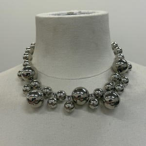 May include: A silver-toned necklace with multiple sizes of round, reflective beads. The beads are strung together, creating a statement piece. The necklace is displayed on a white mannequin.
