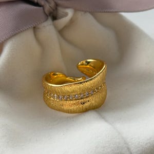 Puede incluir: Un anillo ajustable de color dorado con una superficie texturizada y una fila de pequeñas piedras transparentes. El anillo está abierto en la parte superior y se encuentra sobre una superficie blanca.