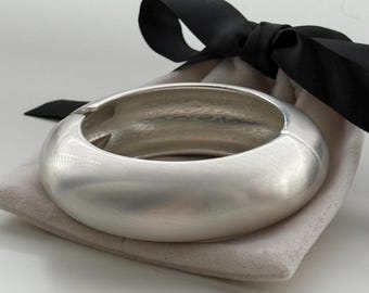 May include: A wide, silver-toned bangle bracelet with a brushed metal finish. The bracelet is resting on a small, light-colored pouch, tied with a black satin ribbon. The bracelet has a smooth, rounded design.