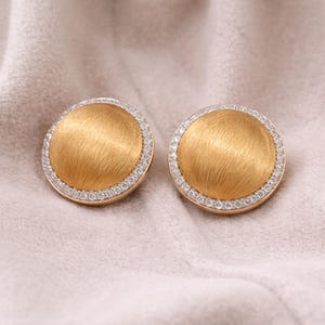 Peut inclure: Une paire de boucles d'oreilles à clous dorées. Chaque boucle d'oreille présente un centre doré brossé entouré d'un anneau de petites pierres précieuses claires. Les boucles d'oreilles sont rondes et reposent sur une surface douce et claire.