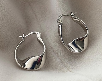 May include: A pair of polished silver hoop earrings. The earrings have a unique twisted design, reflecting light. The metal is bright and shiny, with a smooth surface. The earrings are displayed on a soft, light-colored background.