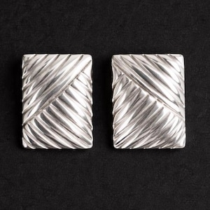 Może przedstawiać: Dwa srebrne, prostokątne kolczyki z teksturowanym, ukośnym wzorem. Kolczyki są prezentowane na ciemnym tle, podkreślając ich metaliczny połysk i skomplikowany design. Kolczyki do przekłutych uszu.