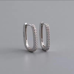 May include: A pair of silver oval hoop earrings. Each earring is set with small, clear stones. The earrings are shown against a grey background. The text "S925" is visible on one earring.