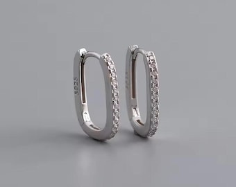 May include: A pair of silver oval hoop earrings. Each earring is lined with small, clear stones. The earrings are displayed on a gray background. The text "S925" is visible on one earring.