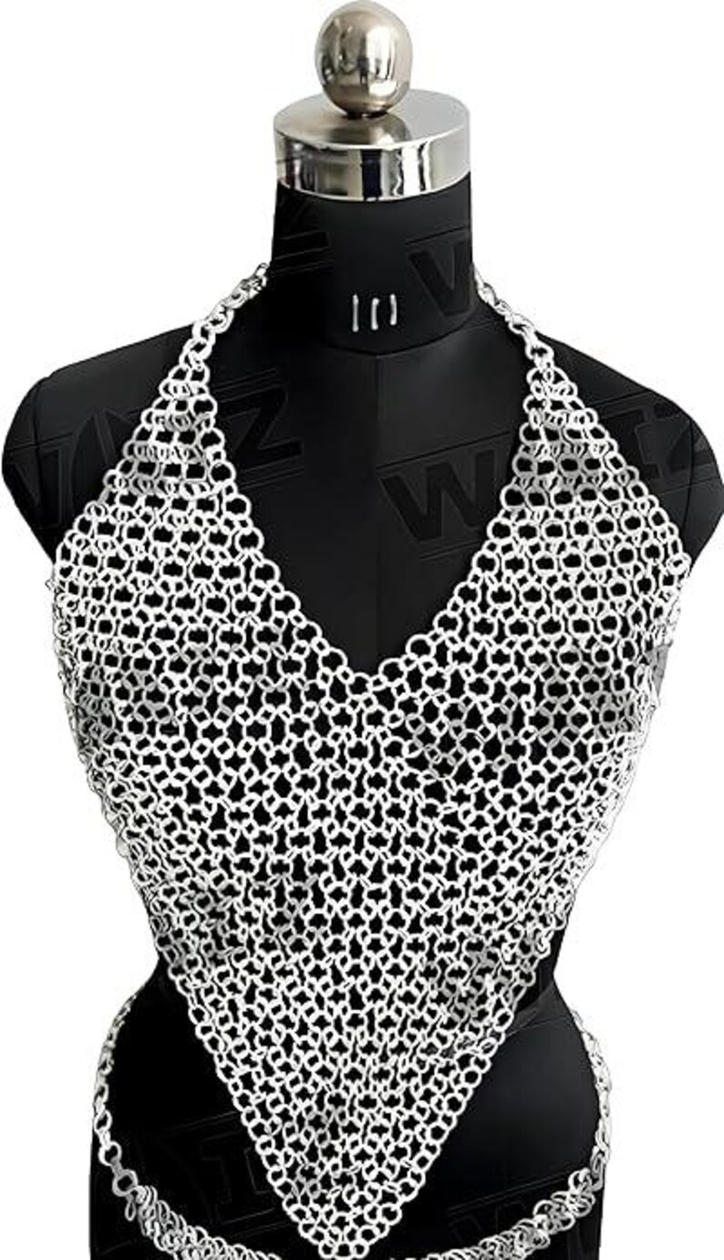 May include: A silver-colored chainmail halter top displayed on a black mannequin. The top features a V-neck design and a matching chain that wraps around the waist. The metal links are intricately woven, creating a mesh-like pattern.