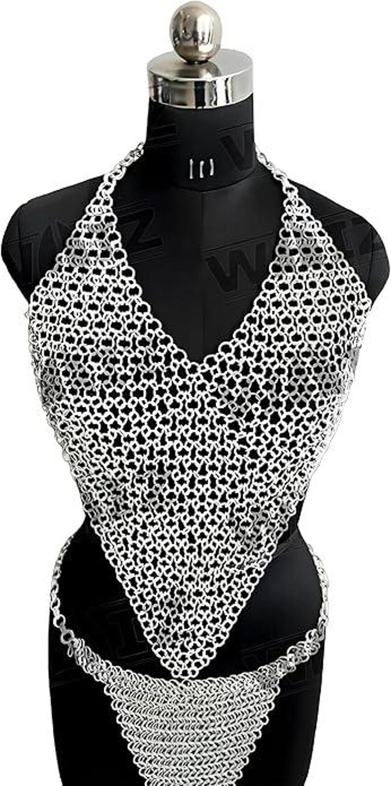 May include: A silver chainmail halter top and matching bottom set displayed on a black mannequin. The chainmail is made of small, interlocking metal rings, creating a textured, open weave design. The top has a deep V-neck.