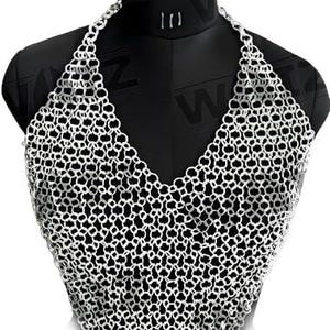 May include: A silver-colored chainmail halter top displayed on a black mannequin. The chainmail is made of small, interlocking metal rings, creating a textured, open weave. The top has a V-neck design and a triangular shape.