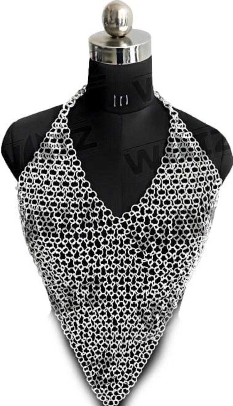 May include: A silver-colored chainmail halter top displayed on a black mannequin. The chainmail is made of small, interlocking metal rings, creating a textured, open weave. The top has a V-neck design and a triangular shape.