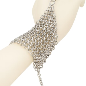 Puede incluir: Pulsera de cota de malla plateada con un accesorio para el dedo. La pulsera está hecha de pequeños anillos de metal entrelazados, formando una superficie flexible y texturizada. Una cadena se extiende desde la pulsera para el cierre. La joya se muestra en una mano de maniquí.