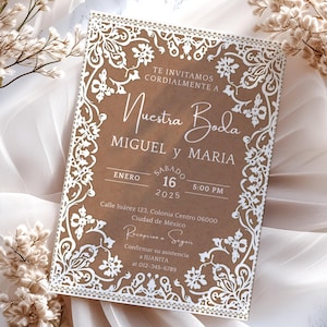 Op de afbeelding: Een trouwuitnodiging met een bruine achtergrond en een wit bloemen kanten ontwerp. De tekst luidt "Nuestra Boda" met de namen Miguel en Maria, samen met de datum, tijd en locatie. De uitnodiging is omgeven door witte bloemen en stof.